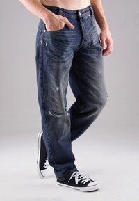 Dunkelblaue Jeans mit verwitterten Stellen, geradem Bein-Schnitt, fünf Taschen und Metallknopfverschluss. Kombiniert mit schwarzen Sneakers im Low-Top-Design.