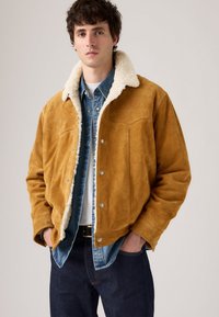 Bruine leren jas van suede met een witte sherpa kraag, gecombineerd met een denim overhemd en donkere jeans. Voorzien van drukknoopsluitingen en decoratieve stiksels.