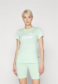 Zeleno-mentolové tričko s krátkym rukávom vyrobené z mäkkej tkaniny, s veľkým bielym logom "PUMA" a siluetou pumy. Skombinované s zodpovedajúcimi mentolovými šortkami.