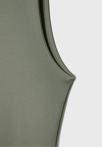 Gros plan sur un haut sans manches vert olive montrant un tissu lisse et des coutures arrondies aux emmanchures sur un fond uni clair.