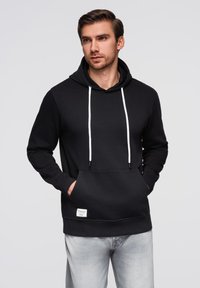 Černá mikina s kapucí a nastavitelnými š�ňůrkami, přední klokaní kapsou a malou nášivkou. Materiál se zdá být hladký a mírně pružný.