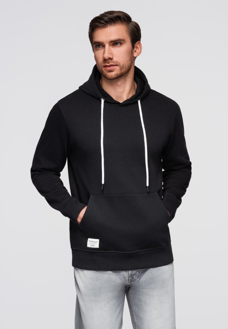 Černá mikina s kapucí a nastavitelnými šňůrkami, přední klokaní kapsou a malou nášivkou. Materiál se zdá být hladký a mírně pružný.