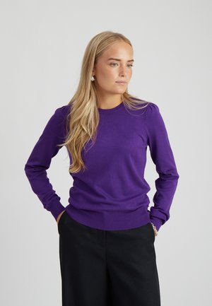 Mujer con cabello largo y rubio que lleva un suéter morado de manga larga y pantalones negros, de pie con las manos en los bolsillos, mirando hacia un lado.