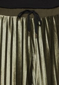 Pantaloni plissettati in velluto olive con cintura a lacci, caratterizzati da strisce verticali, una texture morbida e dettagli in cordoncino nero a contrasto.