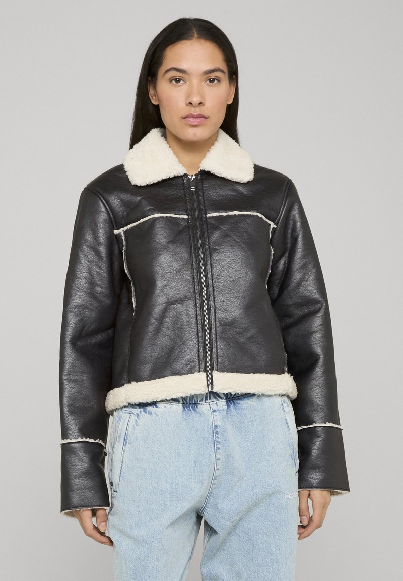 Giacca di pelle nera con collo e orlo in ecopelliccia color crema, con chiusura a zip e dettagli di cuciture visibili. Indossata sopra un denim chiaro.