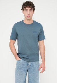 Μπλε βαμβακερό T-shirt με κοντά μανίκια και στρογγυλή λαιμόκοψη, με μικρό εκτυπωμένο λογότυπο στο αριστερό στήθος. Συνδυάζεται με ανοιχτό μπλε τζιν παντελόνια.
