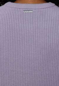 Maglione a coste di un leggero colore viola con scollatura rotonda e una piccola etichetta metallica del marchio sul retro. Tessuto testurizzato con un motivo di cucitura visibile.