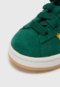 Zöld velúr sneaker, fehér gumitalppal, bézs aljjal, kerek orral és texturált fűzőkkel a stabil illeszkedés érdekében.