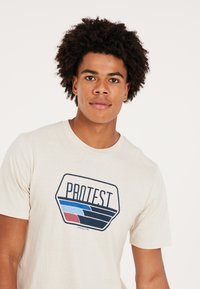 Beige bomulds T-shirt med et blåt og rødt grafisk print med teksten "PROTEST". Kortærmet og design med rund hals.