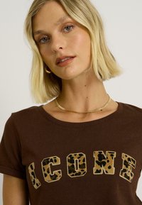 Camiseta de algodón marrón de manga corta y cuello redondo. Presenta letras doradas y con estampado de leopardo que dicen "ICON". Textura suave.