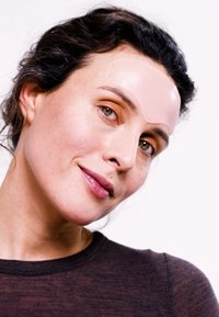 Durchsichtiger Gel-Pflaster auf der Stirn, helle Hautfarbe, kurzes dunkles welliges Haar, trägt ein dunkles Oberteil, dezentes natürliches Make-up, neutraler Hintergrund.