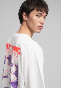 T-shirt oversize blanche avec un imprimé graphique coloré au dos, comprenant des éléments rouges et violets, fabriquée en tissu de coton doux.