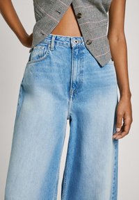 Jeans de pierna ancha en azul claro con una textura desgastada, que cuentan con cinco bolsillos y costuras visibles. La cintura tiene un cierre de botón.