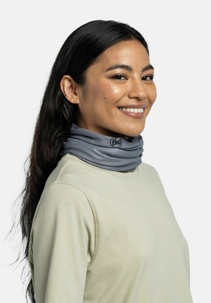 Frau mit langen dunklen Haaren, die ein beiges Top und einen grauen Buff-Neckwarmer trägt, lächelt und schaut leicht zur Seite.