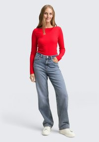 Rotes geripptes Langarmoberteil, kombiniert mit hellblauen hoch taillierten weit geschnittenen Jeans. Das Modell trägt weiße Sneaker. Einfaches, lässiges Outfit.