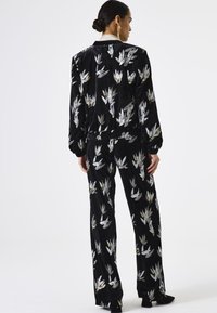 Zwarte fluwelen outfit bestaande uit een bomberjack en bijpassende broek. Versierd met metalen bloemenpatronen en elastische manchetten.