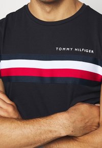 Mustapaita, jossa on lyhyet hihat ja vaakasuora raitakuvio punaisena, valkoisena ja tummansinisenä. Paidan yläosassa on "TOMMY HILFIGER" -teksti raidoituksen yläpuolella.