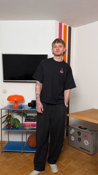 Ung mann med tatoveringer står i et rom med en TV på veggen, oransje lampe, basketball, bøker og en metallisk Tropicana-koffert.