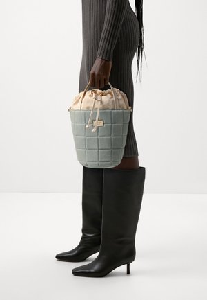 Grå quiltet bøtte-taske med beige snor i toppen, holdt af en hånd. Har guldfarvet hardware og et rektangulært mærke. Sorte knælange støvler er synlige.