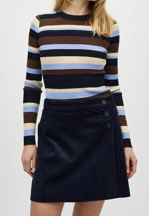 Kobieta ubrana w prążkowany sweter z długim rękawem w brązowe, kremowe, niebieskie i czarne paski, w połączeniu z granatową spódnicą sztruksową z trzema guzikami.