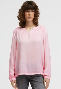 Frau mit lockigem braunem Haar, die eine hellrosa Bluse mit langen Ärmeln und dunkelgraue Jeans trägt, steht vor einem schlichten weißen Hintergrund.