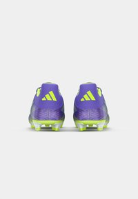 Des crampons de football violets et vert lime avec une surface texturée, présentant un design élégant et un talon renforcé, mis en valeur par un branding bien en vue.