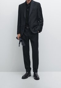 Czarny garnitur z dopasowanymi spodniami i przylegającą czarną koszulką polo. Błyszczące czarne skórzane buty. Krój slim-fit, minimalna ilość metalowych elementów, gładka faktura.