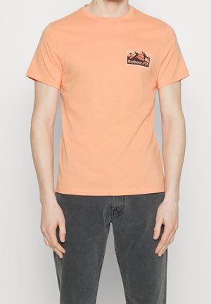 Mężczyzna w brzoskwiniowej koszulce z krótkim rękawem z małą grafiką góry i tekstem "Barbour" na górnej lewej stronie klatki piersiowej, w połączeniu z szarymi spodniami.