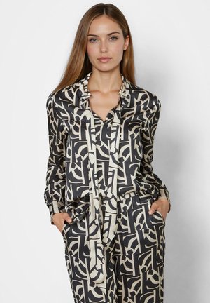 Woman with long straight hair wearing a black and beige abstract patterned blouse and matching pants, hands in pockets, neutral background.