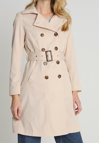 Trench coat beige con diseño de doble botonadura, cinturón ajustable, acentos de botones y bolsillos laterales, fabricado en una tela suave.