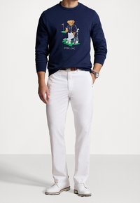 Marinblå sweatshirt med ett björngrafik som håller golfklubbor, vita byxor och vita skor. Sweatshirten har rund hals och ribbade muddar.