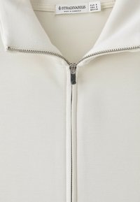 Gros plan d’un vêtement blanc avec fermeture éclair et col, affichant une étiquette Stradivarius et les détails des tailles EUR 8, USA 8, MEX 26.
