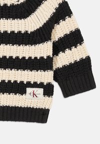 Črtasta pulover v črni in kremni barvi, pletena tekstura, z širokimi vodoravnimi črtami, rebrastimi manšetami in narobe obrnjenim logotipom Calvin Klein Jeans.