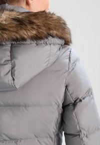 Giubbotto imbottito grigio con design trapuntato, dotato di bordo del cappuccio in pelliccia sintetica e texture liscia e lucida. Nessun logo o motivo visibile.