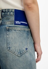 Gaiši zili džinsi ir aprīkoti ar lielu zilu ādas uzšuvi, kurā baltā krāsā ir iespiests "KARL LAGERFELD JEANS", un neliels zils logotips aizmugurē džinsu kabatā.