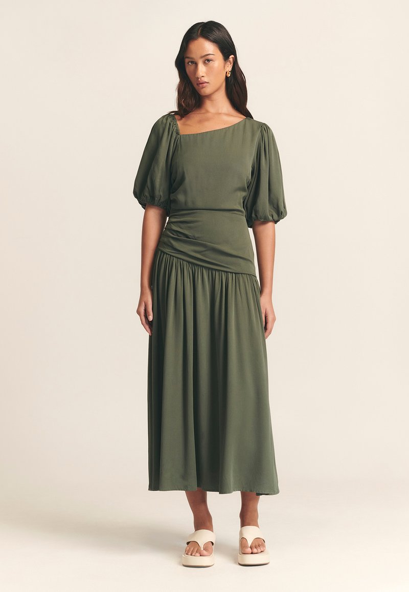 Olivgroene jurk van middelbare lengte met pofmouwen, geplooide taille en geplooide rok. Draagt crèmekleurige plateausandalen. Glad stoftextuur.