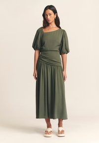 Abito midi verde oliva con maniche a sbuffo, corpino aderente e gonna arricciata. Tessuto dalla trama liscia. Indossato con sandali platform beige.