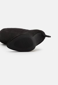 Botte haute jusqu'au genou en daim noir avec un bout pointu, un talon fin et une semelle en caoutchouc texturée avec des rainures horizontales.