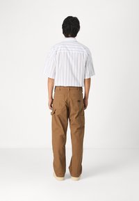 Bruna cargo-byxor med flera fickor, kombinerat med en kortärmad vit skjorta med smala marinblå ränder. Modell vänd mot ryggen.