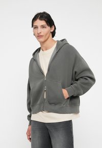 Harmaa vetoketjuhuppari pehmeästä materiaalista, jossa on etutasku, säädettävä huppu ja rento istuvuus, kerroksittain kevyen paidan päällä.