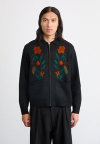 Juodas megztinis su gėlių siuvinėjimu oranžine ir žalia spalvomis, turintis užtrauktuką priekyje, apykaklę ir dvi priekinės kišenes.