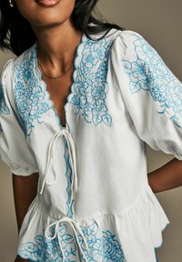 Blusa bianca con raffinati ricami floreali blu lungo il collo e le maniche, con orlo a peplum e laccio frontale.