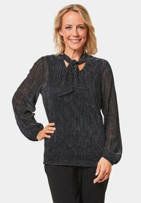 Blouse noire à manches longues avec une finition texturée, dotée d'un nœud au cou, de manches semi-transparentes et d'une coupe droite.