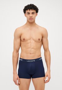Boxers azuis marinhos feitos de uma mistura suave de algodão, com uma ampla cintura elástica estampada em branco com a marca "LACOSTE" e um design ajustado.