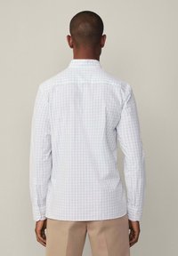 Camicia bianca a maniche lunghe con un leggero motivo a griglia. Presenta un colletto classico e polsini con bottoni. Il tessuto appare liscio e strutturato.