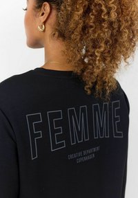 Melns džemperis ar izteiksmīgu vārdu "FEMME" uz muguras treknām burtiem, ar mīkstu auduma tekstūru un brīvu piegriezumu.