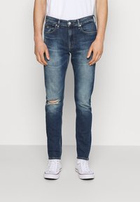 Jeans in denim skinny-fit di colore blu scuro con leggeri sbiadimenti e un ginocchio strappato. Presentano un classico stile a cinque tasche e una timbratura sottile.