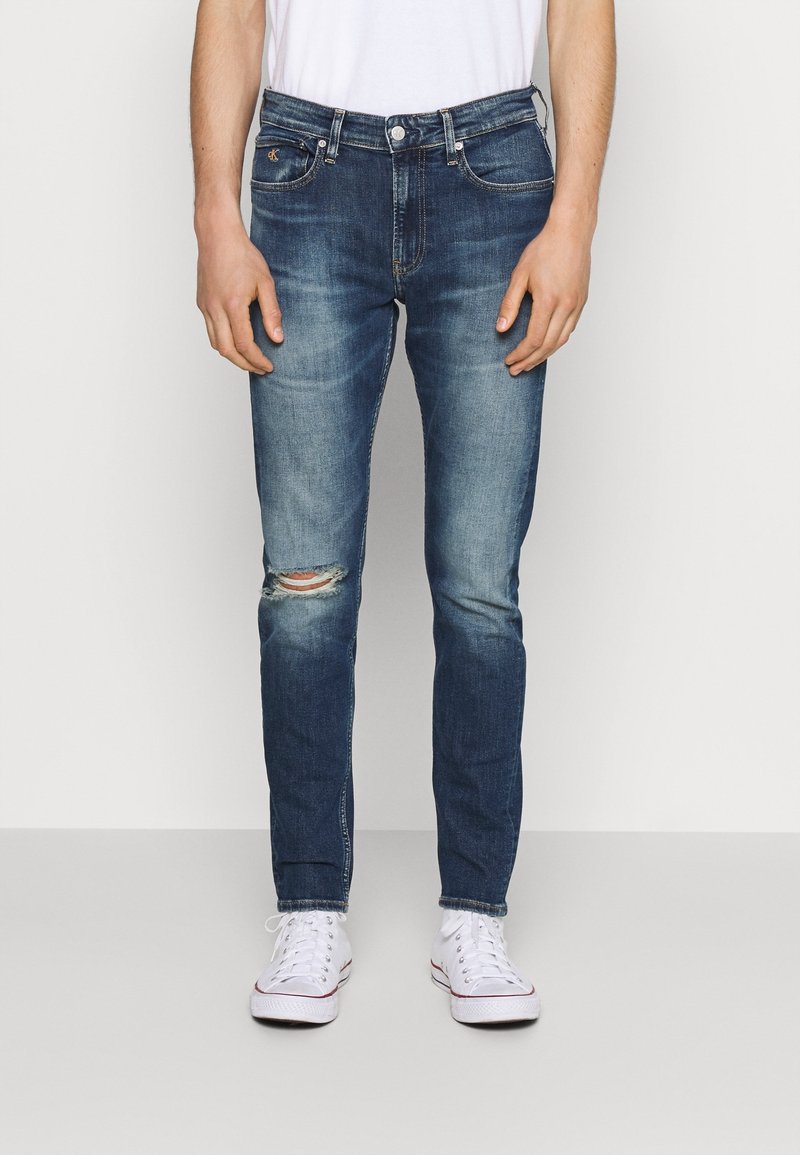 Jeans in denim skinny-fit di colore blu scuro con leggeri sbiadimenti e un ginocchio strappato. Presentano un classico stile a cinque tasche e una timbratura sottile.