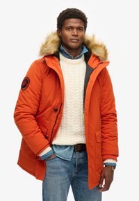Chaqueta de abrigo naranja con capucha con trim de pelo, con cierre de cremallera, textura acolchada y parches negros. Llevada sobre un suéter de punto trenzado.