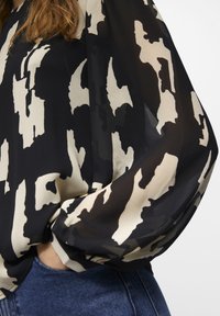 Blusa de color negro y crema con un diseño fluido, que presenta mangas largas y una textura ligeramente translúcida, combinada con jeans de mezclilla azul.